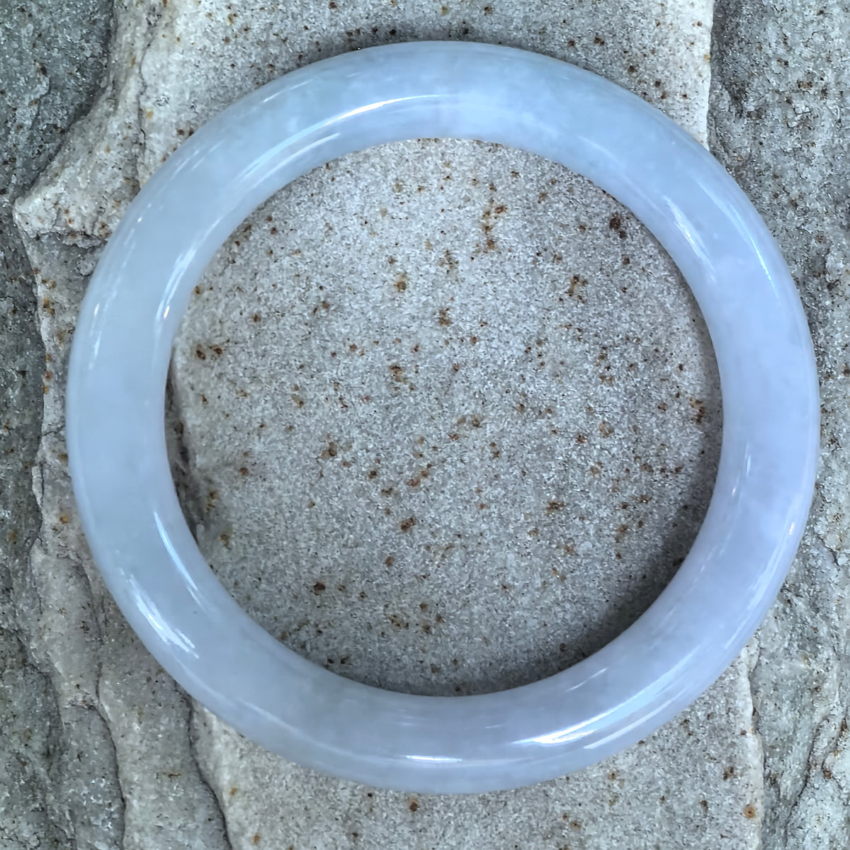 Overhead view of a very pale lavender round-bar jadeite bangle on stone, showing the circular shape and subtle blue-lavender tone.