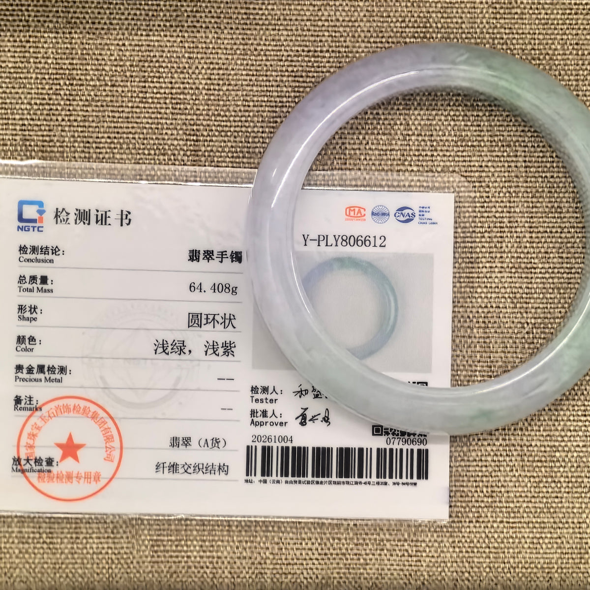 Pale green and lavender jadeite bangle beside its matching NGTC certificate, number Y-PLY806612, on a woven background.