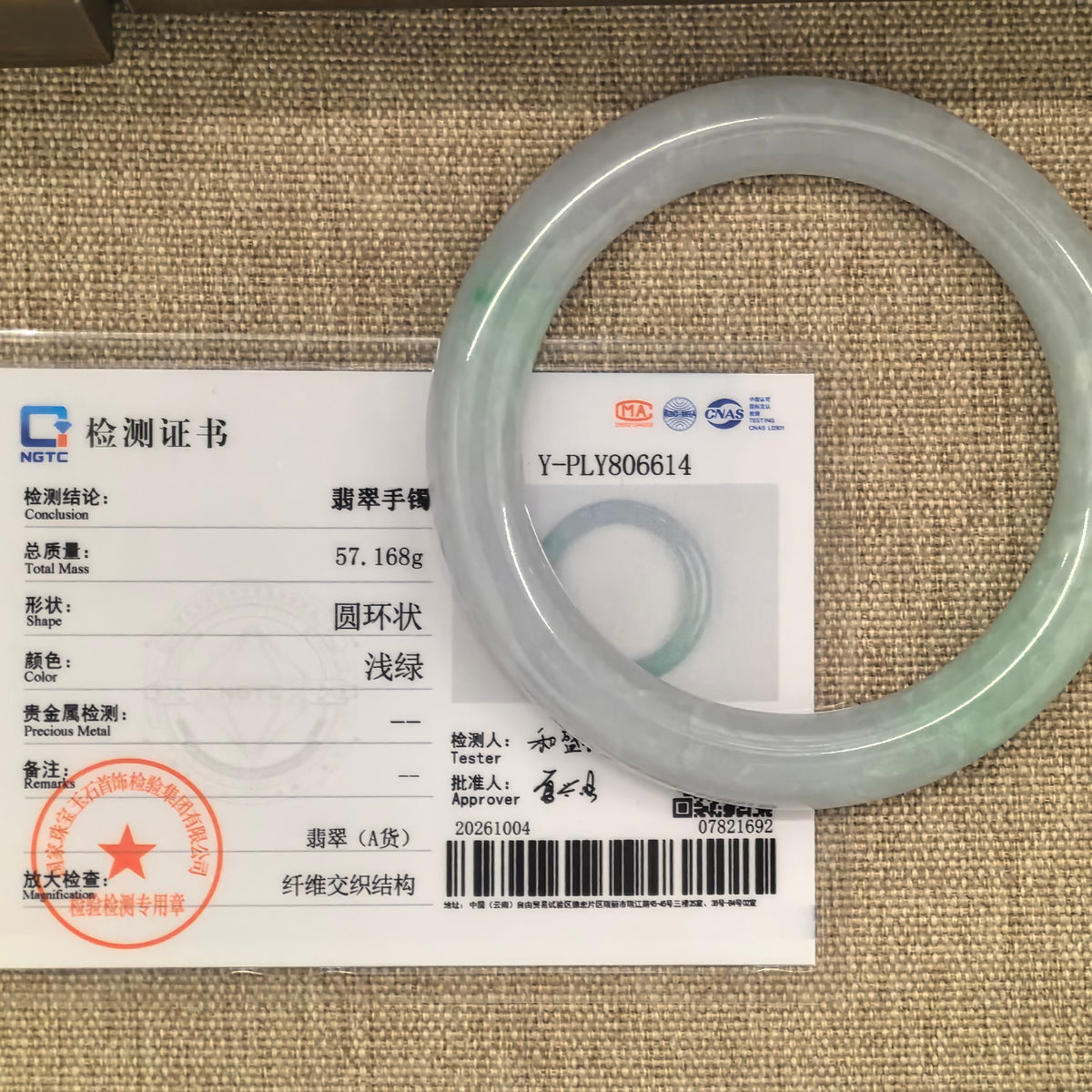 Pale green and pale lavender jadeite bangle beside its matching NGTC certificate, number Y-PLY806614, on a woven background.