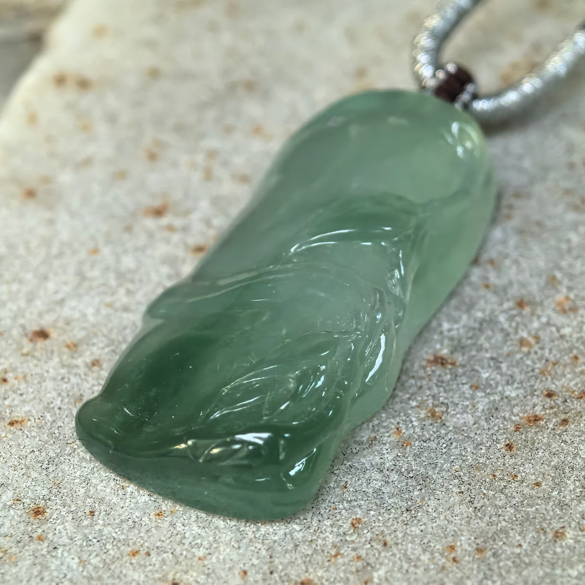 Angled close-up of the icy green jadeite Bamboo Pendant on stone, showing carved leaves, raised bamboo nodes and polished rounded edges.