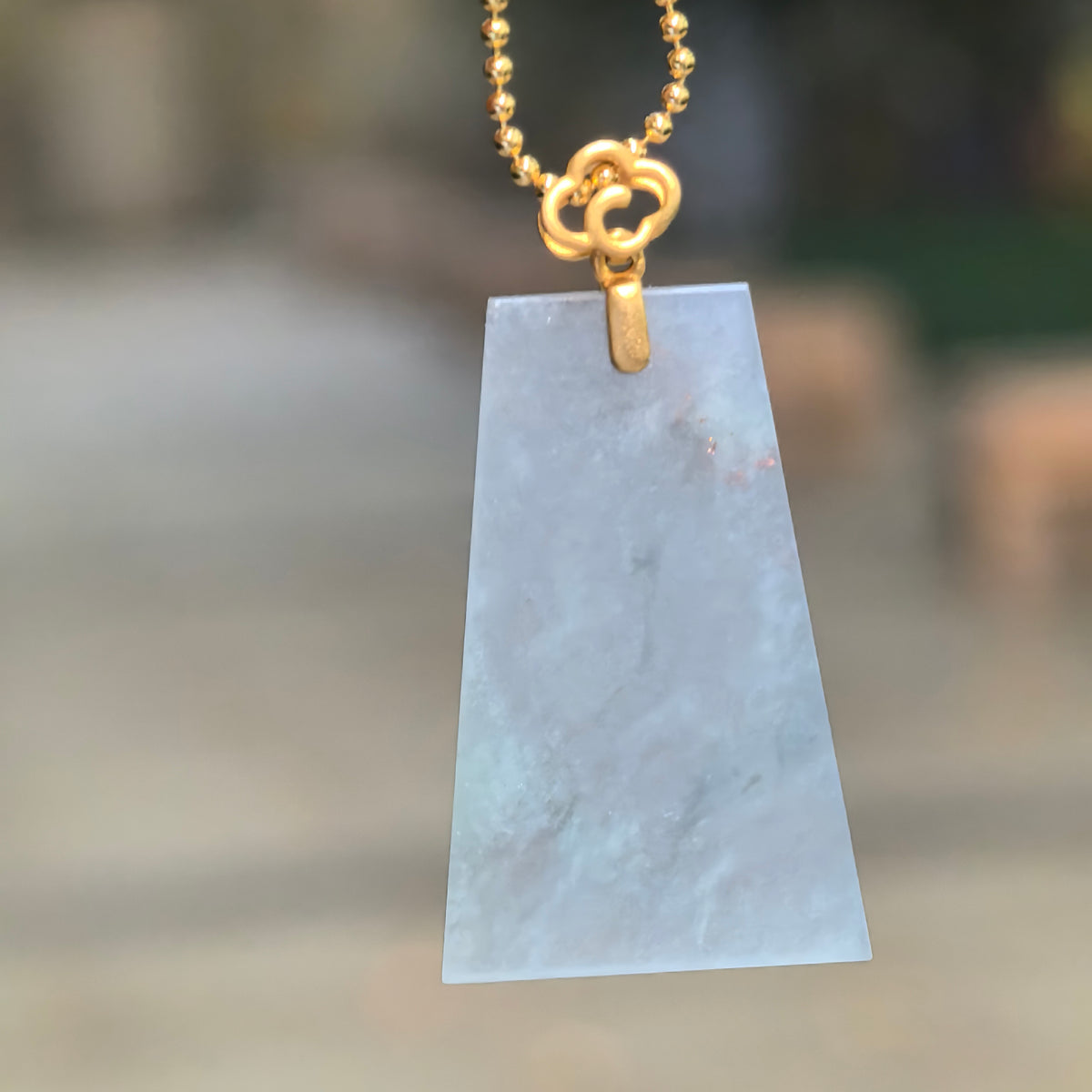 Pale blue jadeite Plain Plaque Pendant hanging from a gold-colored bead chain, showing its trapezoidal shape, translucent body and cotton-like inclusions.