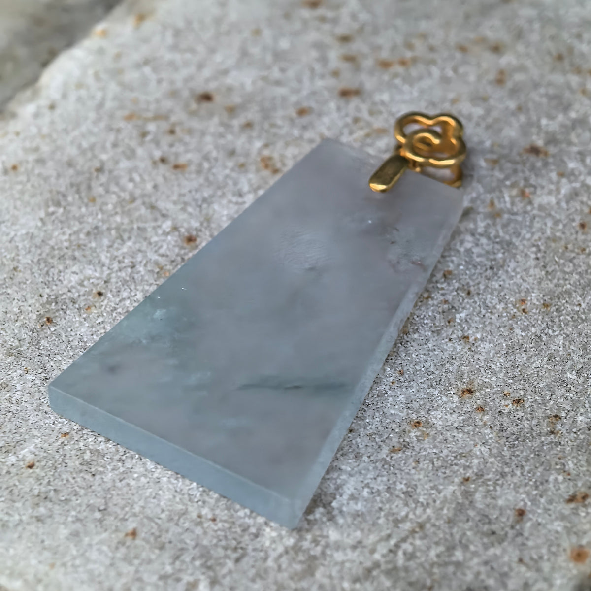 Angled close-up of the pale blue jadeite Plain Plaque Pendant on stone, showing its thin profile, cloudy inclusions and decorative gold bail.