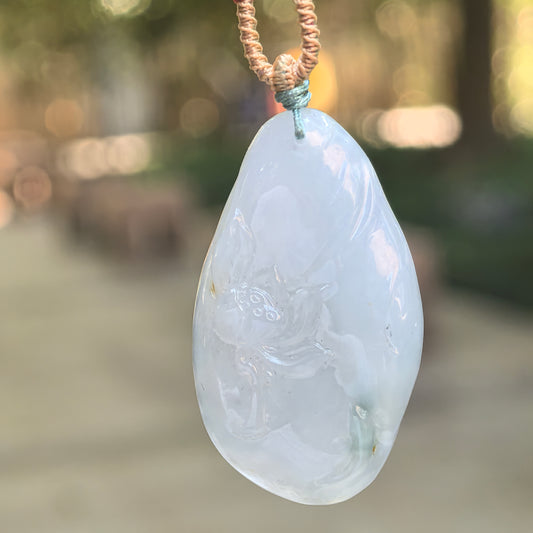 Glutinous white jadeite Lotus and Fish Pendant with a subtle blue undertone, hanging from a knotted cord against a blurred outdoor background.