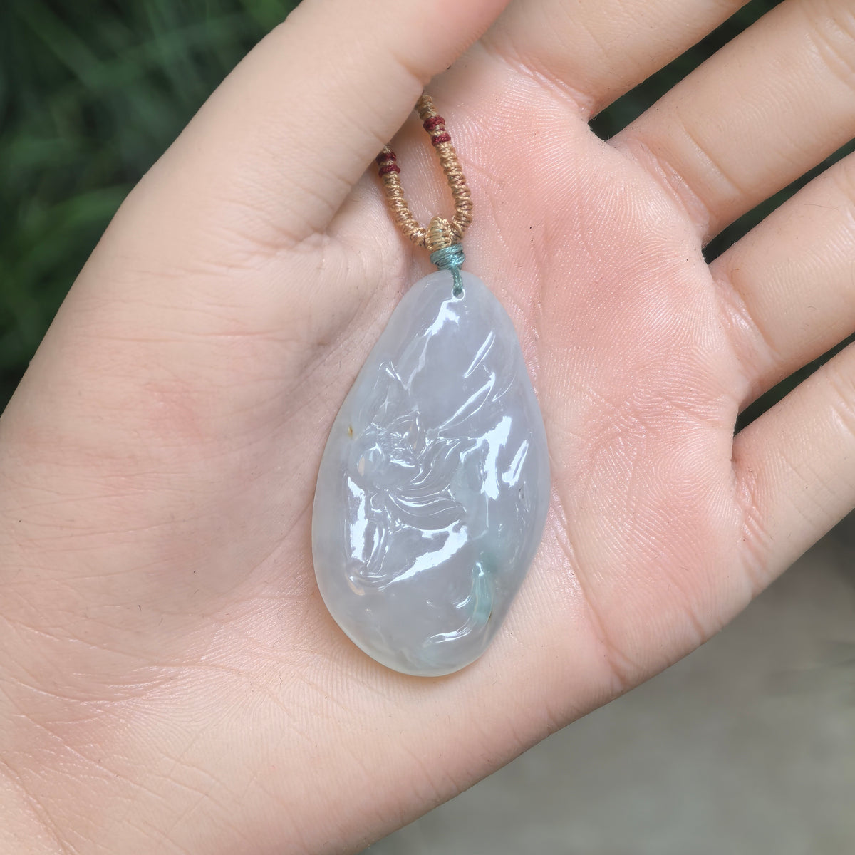 White jadeite Lotus and Fish Pendant with a blue undertone resting in a palm, showing its elongated freeform outline and carved front.