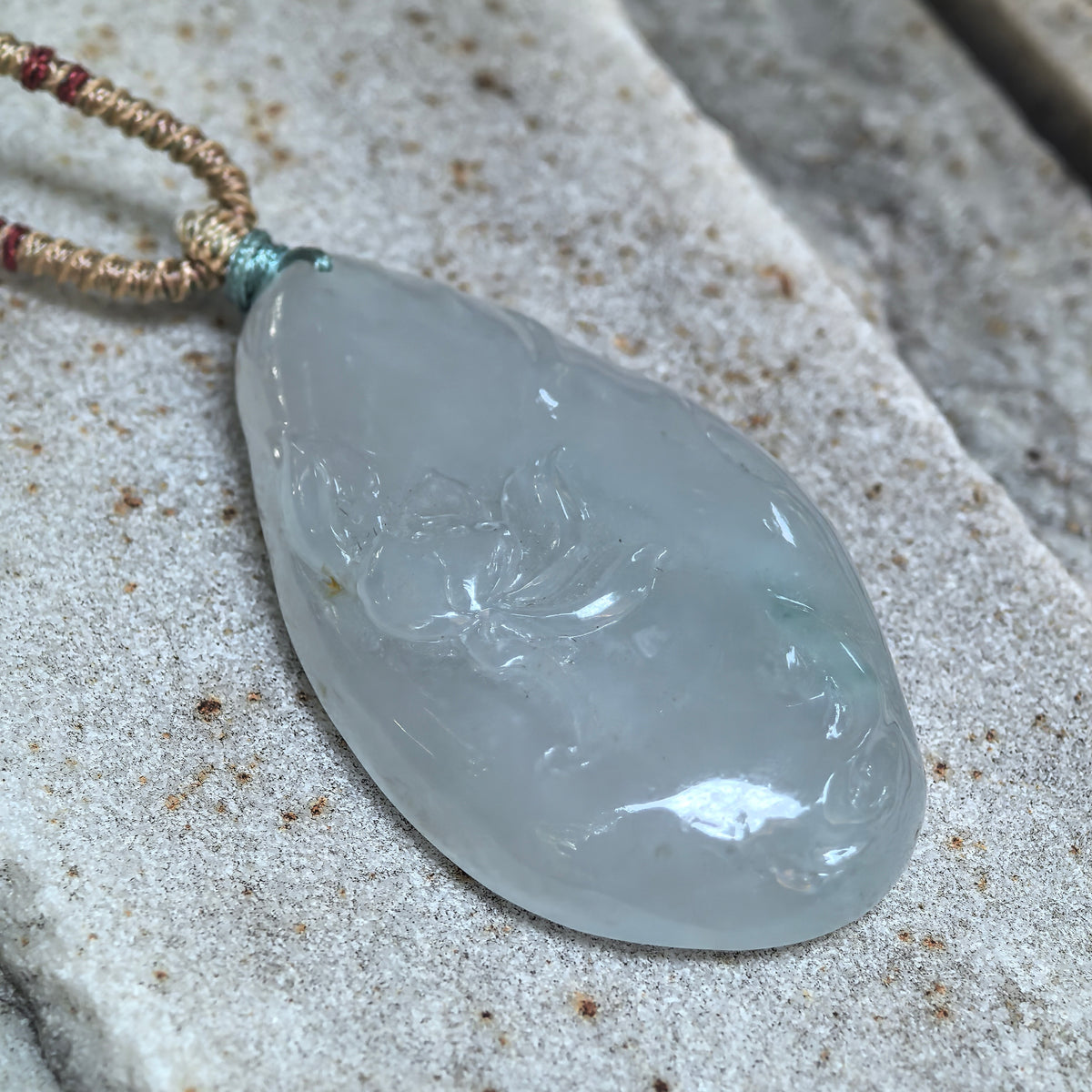 Angled close-up of the white jadeite Lotus and Fish Pendant on stone, showing relief carving, polished edges and a subtle blue undertone.