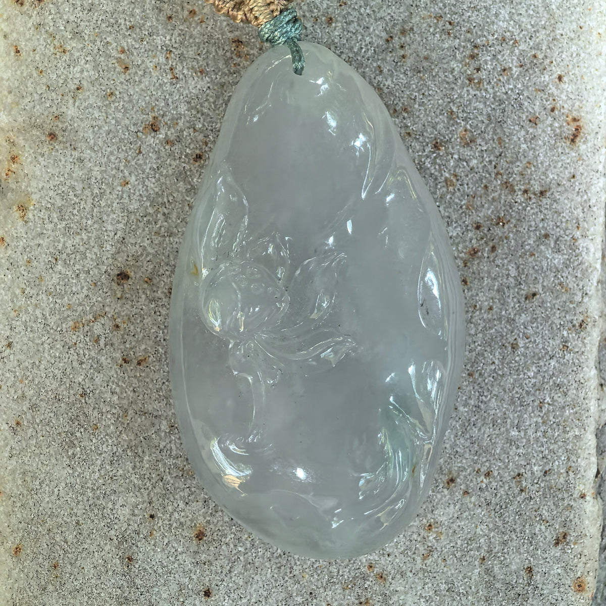 Front view of the white jadeite Lotus and Fish Pendant on stone, showing the broad lotus leaf, flowing fish details and small darker flecks.