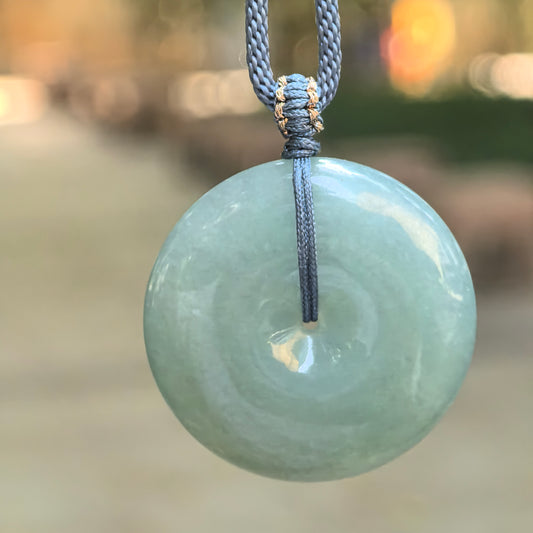 Gray-green jadeite Peace Pendant hanging from a blue-gray cord with gold-colored thread accents against a softly blurred outdoor background.