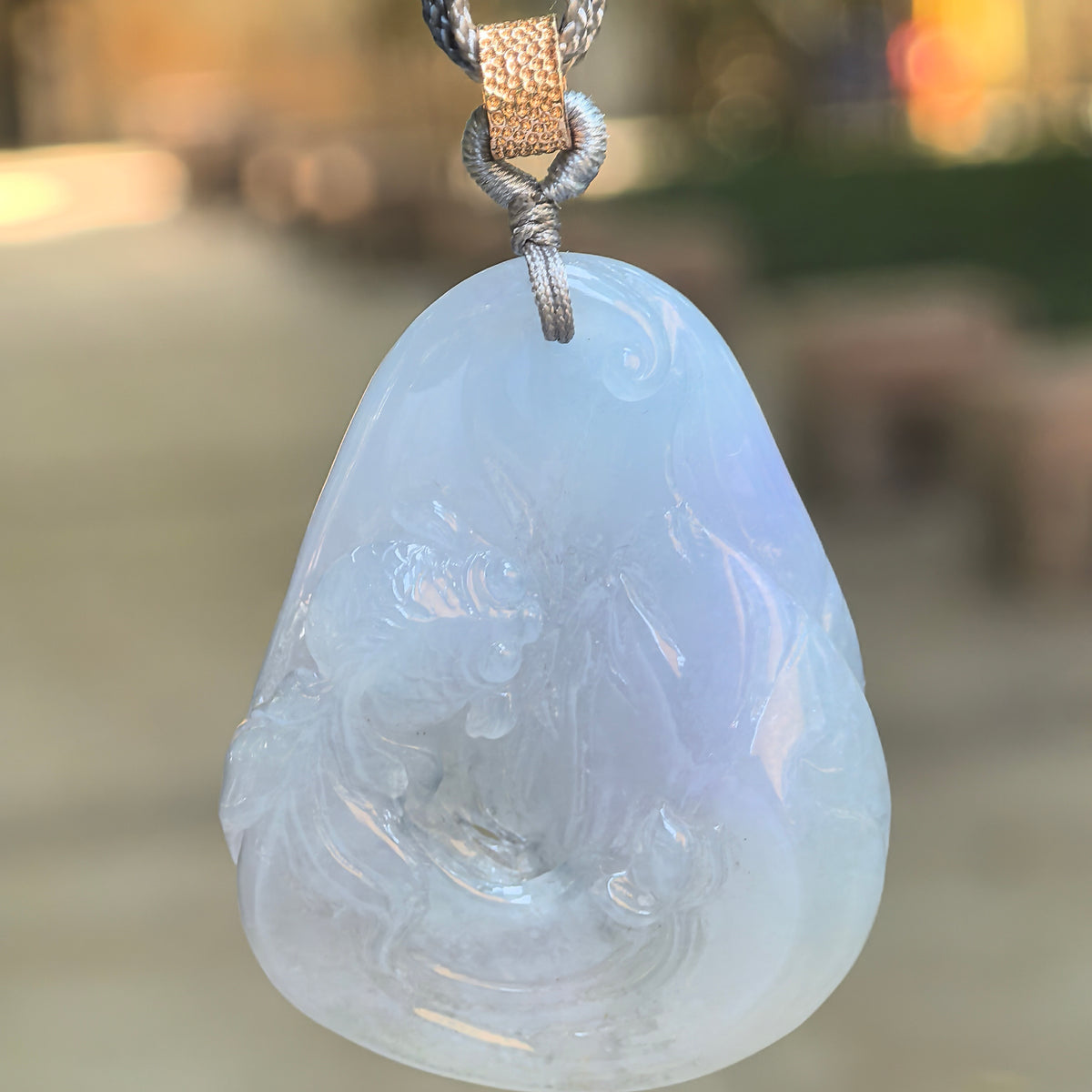 Blue-lavender jadeite Lotus and Fish Pendant hanging from a braided cord against a softly blurred outdoor background.