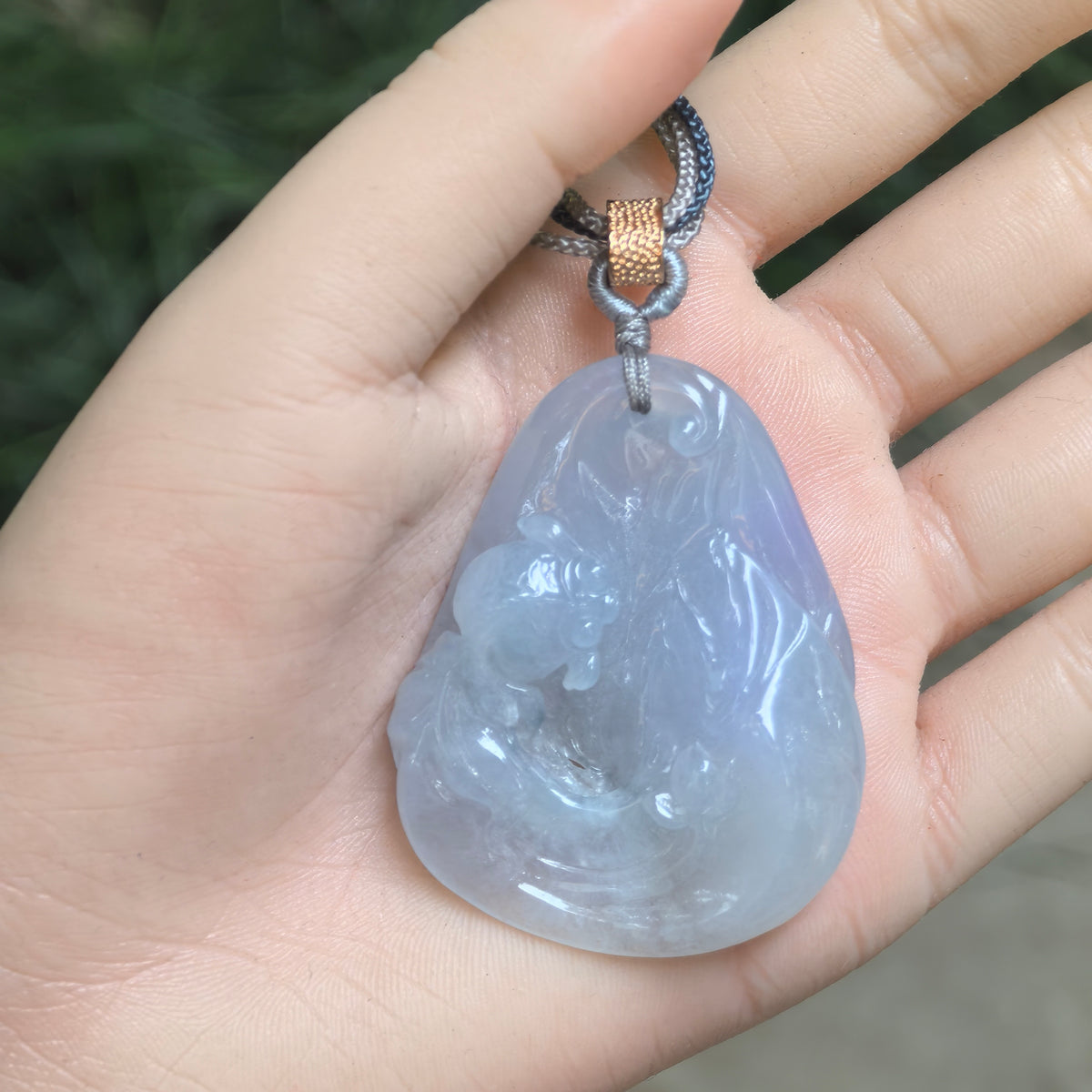 Blue-lavender glutinous jadeite Lotus and Fish Pendant resting in a palm, showing its freeform outline, relief carving and braided cord.