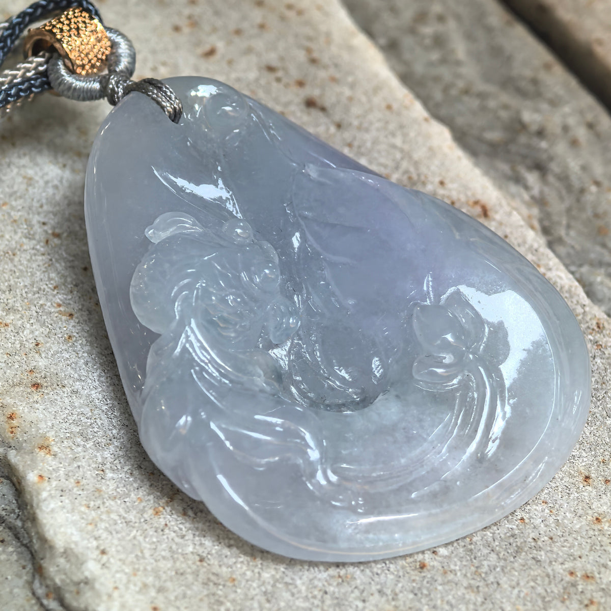 Angled close-up of the blue-lavender jadeite Lotus and Fish Pendant on stone, showing carved fish details, lotus foliage and its rounded edge.