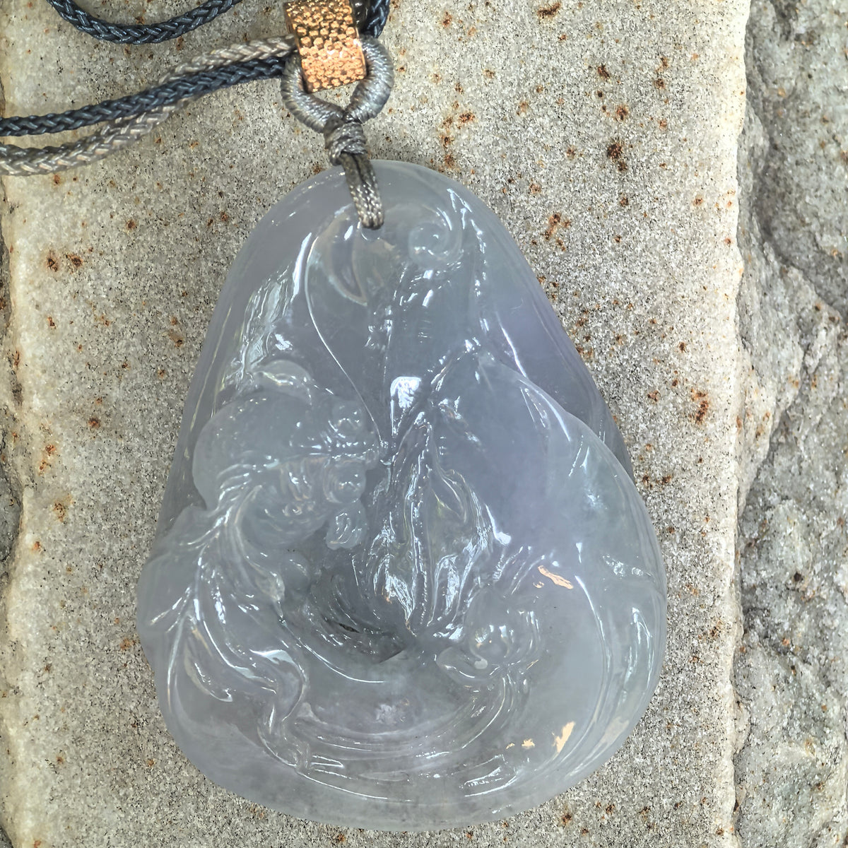Front view of the blue-lavender jadeite Lotus and Fish Pendant on stone, showing flowing relief carving and polished highlights.