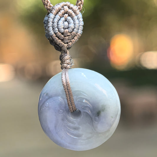 Glutinous lavender jadeite Yin-Yang Fish Peace Pendant hanging from a taupe braided cord, showing paired fish carving and contrasting paler areas.