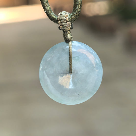 Icy glutinous blue jadeite Peace Pendant hanging from an olive-green knotted cord, showing its translucent circular form against a blurred outdoor background.