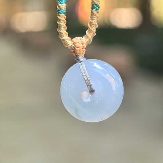 Icy glutinous lavender jadeite Peace Pendant hanging from a gray cord with beige and teal knotted accents against a softly blurred outdoor background.