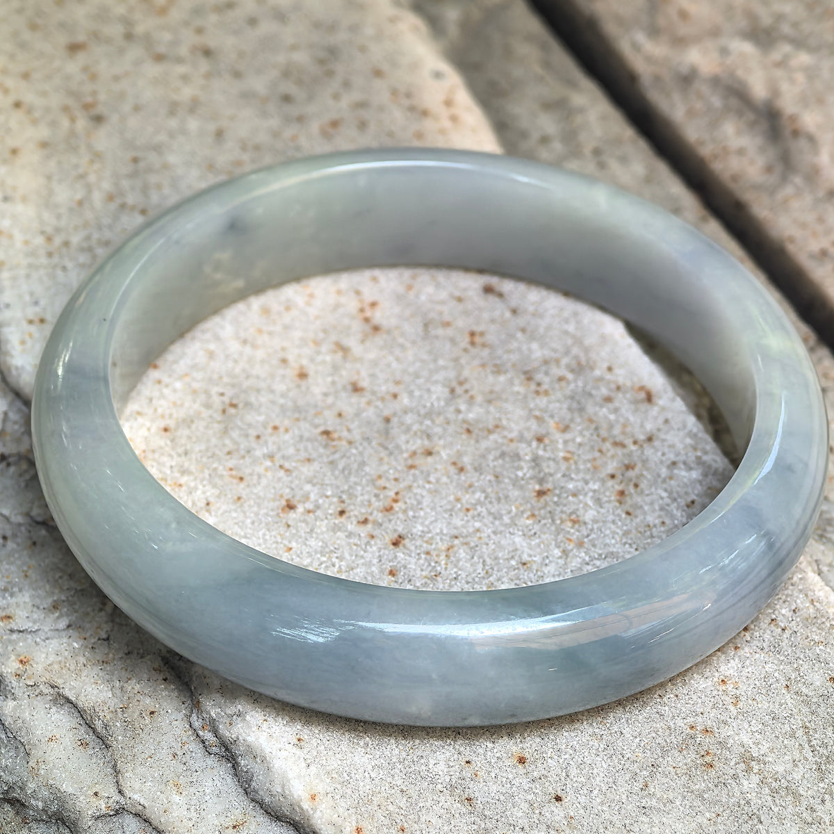 Angled view of a denim blue jadeite bangle resting on stone, showing its floating patterns, rounded outer profile and flatter inner face.
