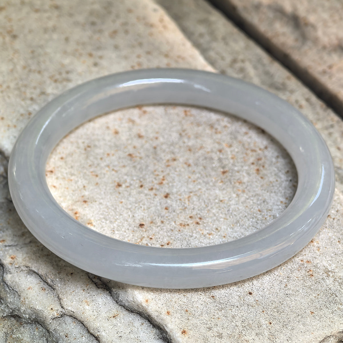 Angled view of a white icy glutinous jadeite round-bar bangle resting on stone, showing its pale green undertone and smooth polished finish.