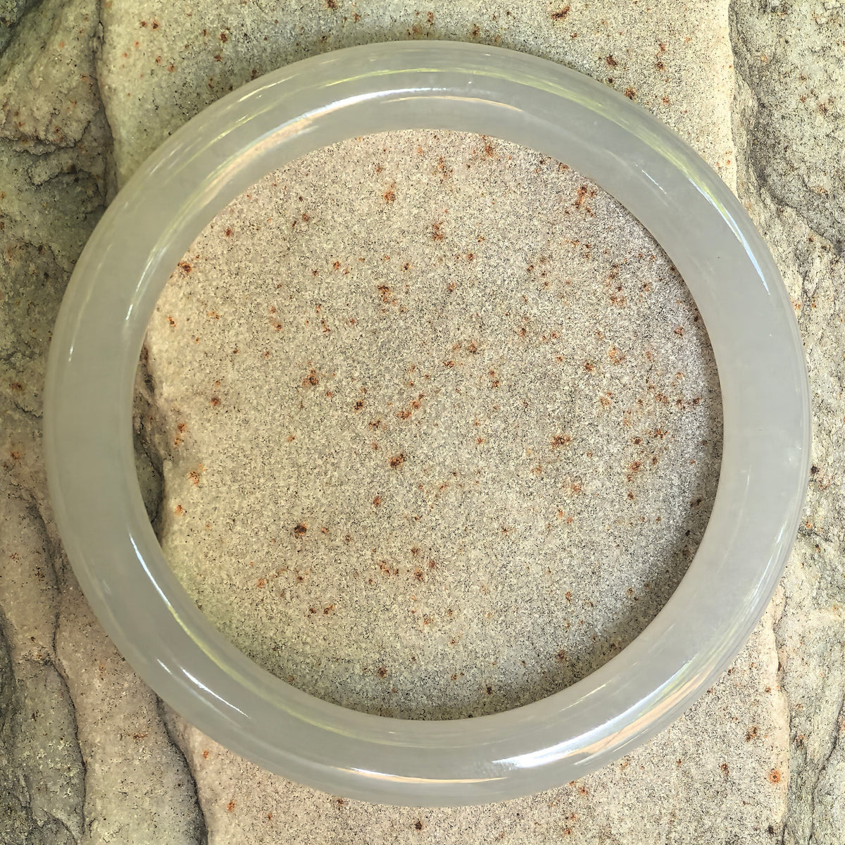 Overhead view of a white jadeite slim round-bar bangle on textured stone, showing its circular outline and subtle pale green tone.