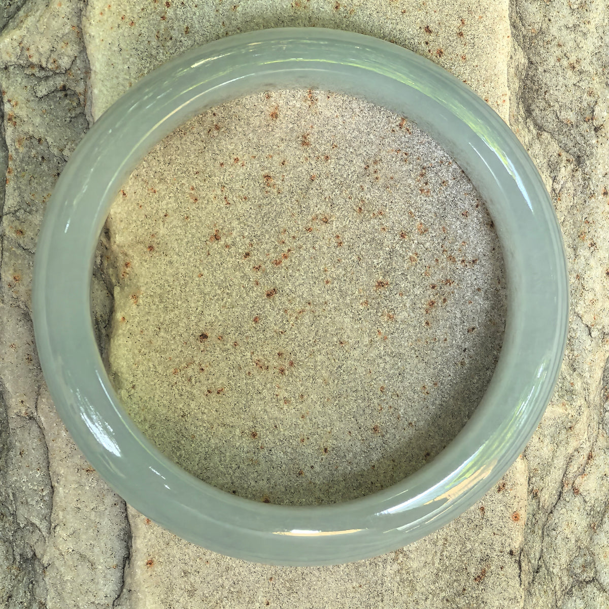 Overhead view of a light green jadeite D-shape bangle on a textured stone surface, showing its circular outline and even overall color.
