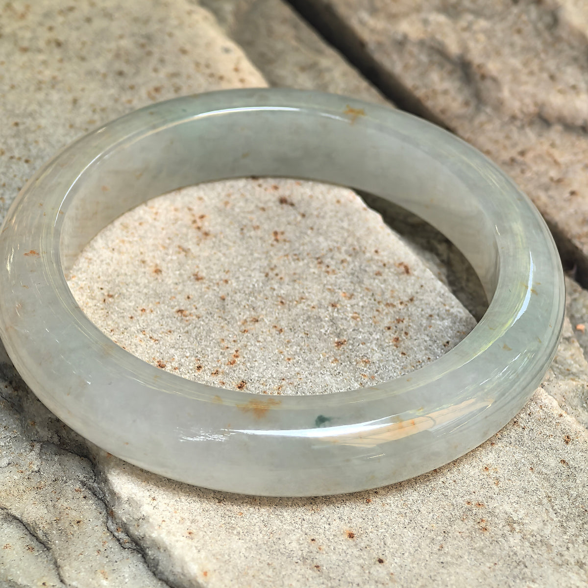 Angled close-up of the pale green D-shape jadeite bangle on stone, showing its flatter inner surface, golden-brown flecks and cotton-like inclusions.