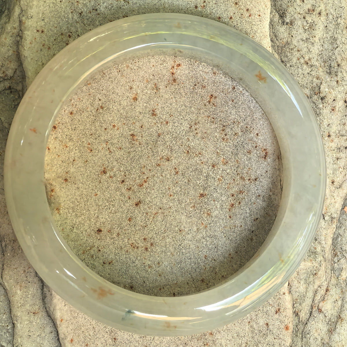 Overhead view of the pale green D-shape jadeite bangle on stone, showing the circular outline and natural golden-brown flecks around the jadeite.