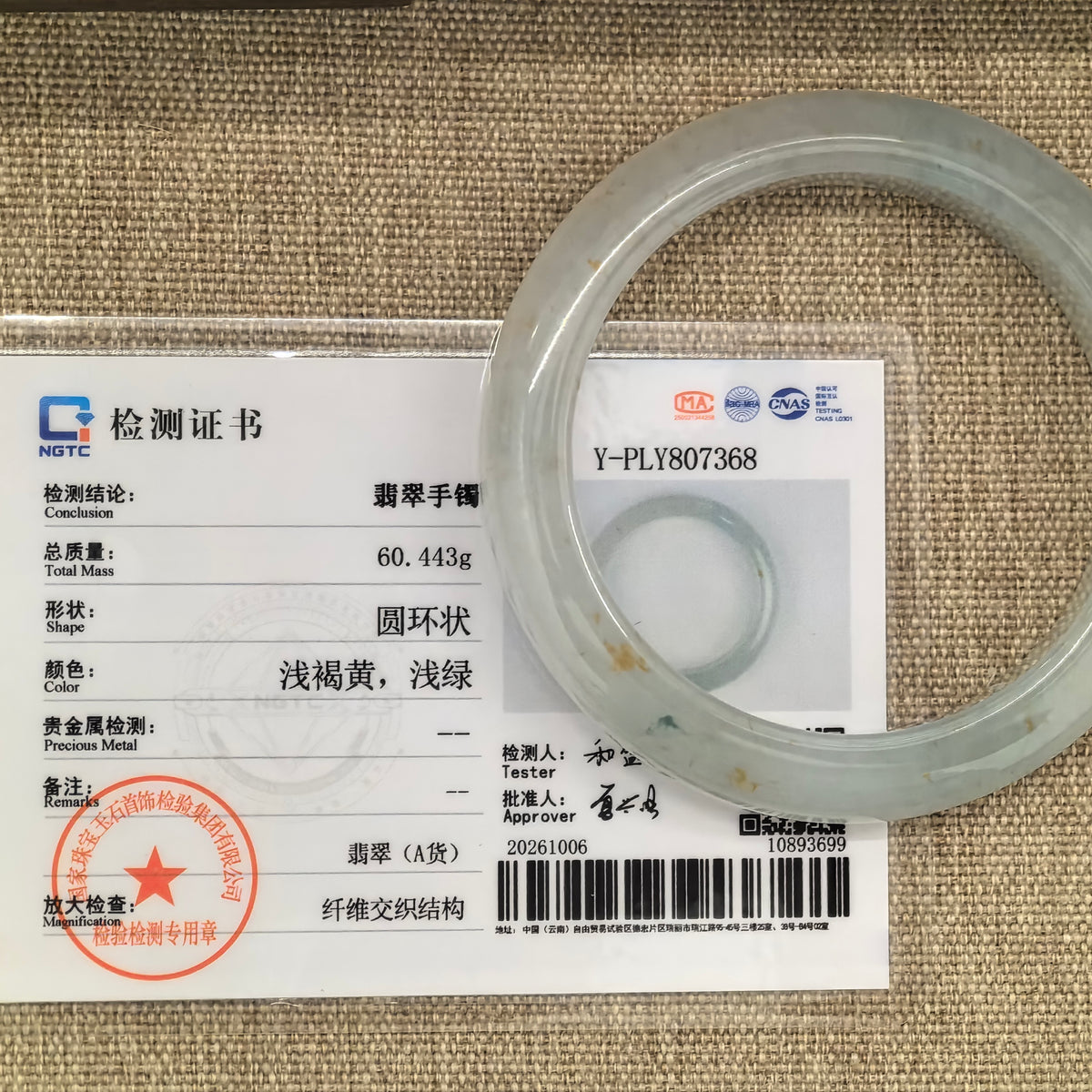 Pale green D-shape jadeite bangle with natural golden-brown flecks beside its matching NGTC certificate.