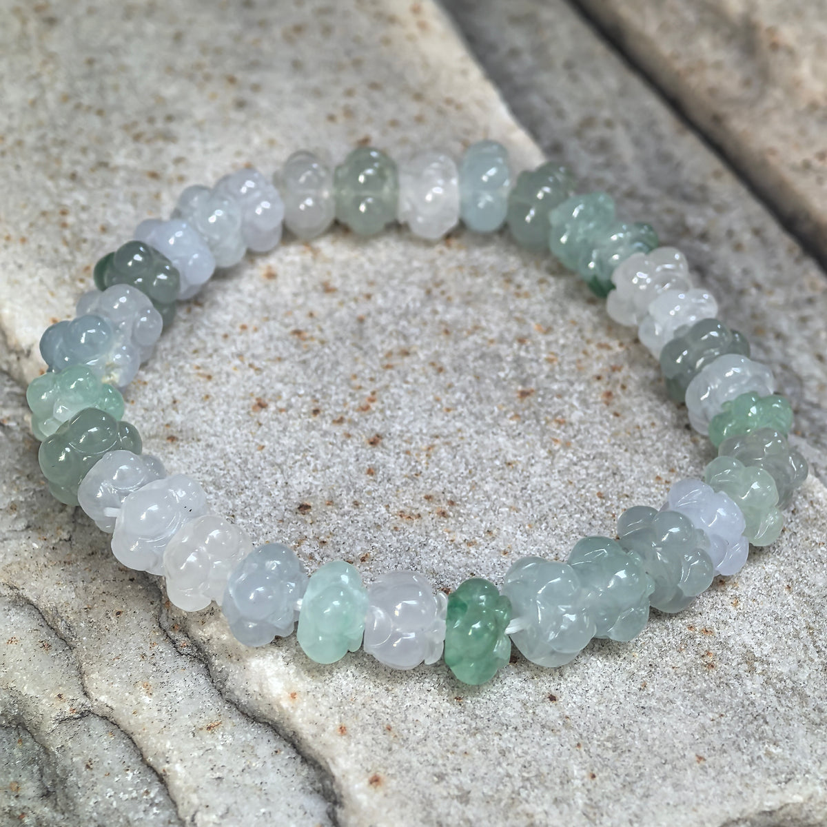 Angled view of a light green and white jadeite lotus bead bracelet arranged on stone, showing the floral carvings around the strand.