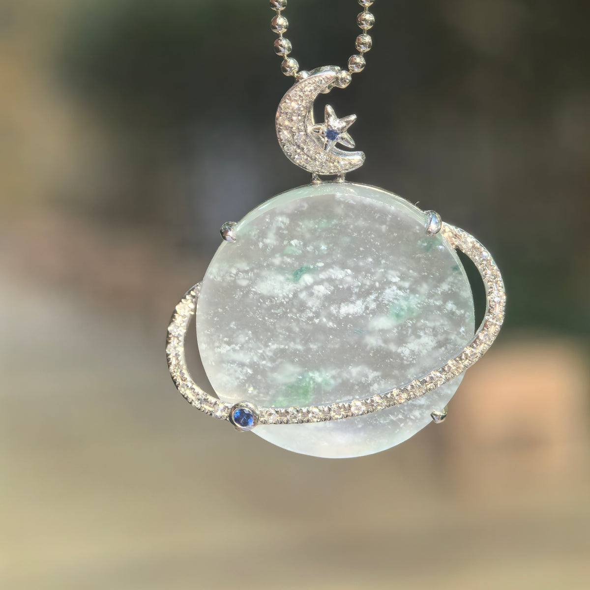 Hanging view of a pale green jadeite Planet Pendant, showing snowflake-like inclusions, green accents, a diamond-set orbital ring and crescent moon and star bail.