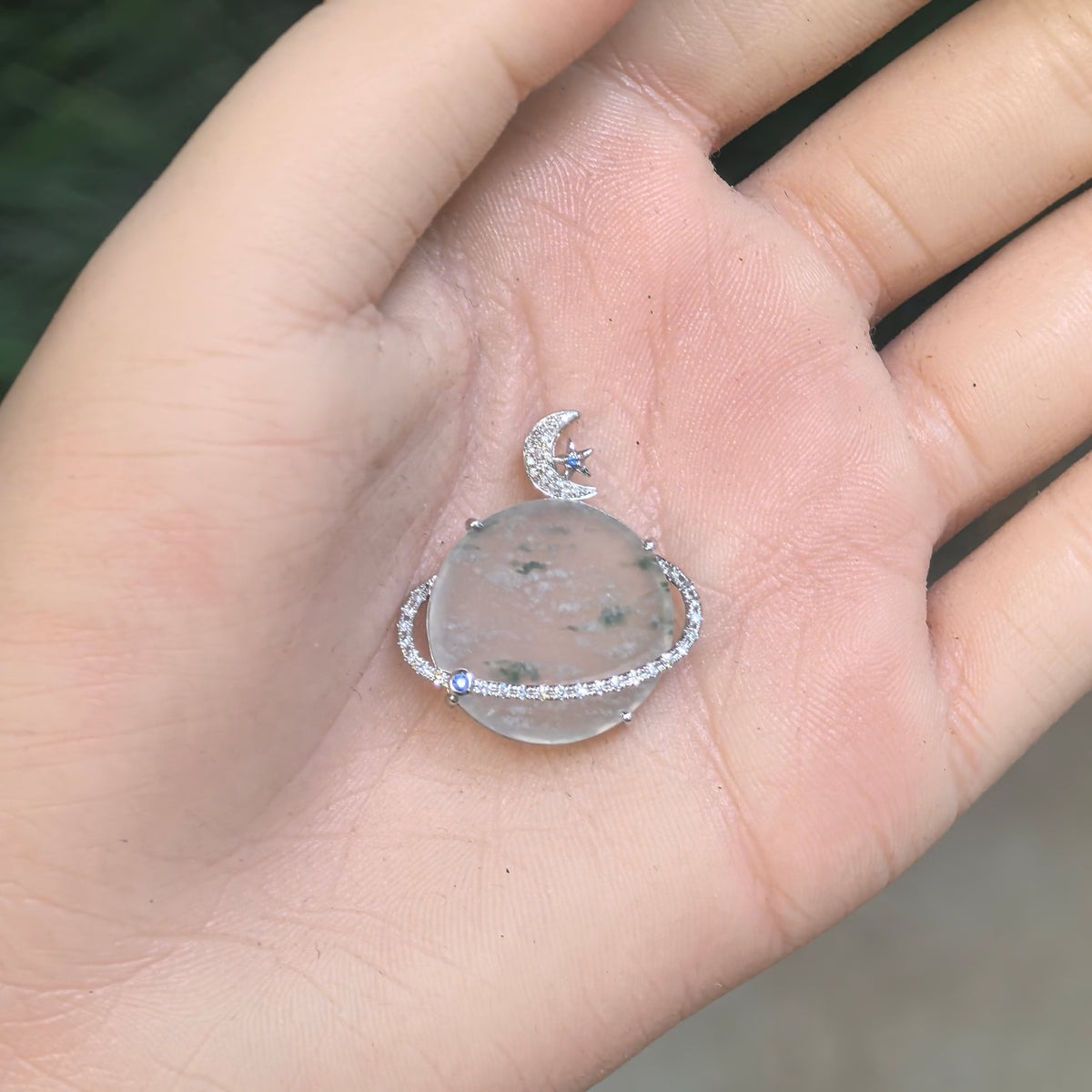 Pale green icy jadeite Planet Pendant resting on a palm, showing its translucency, snowflake-like inclusions and celestial setting with blue accent stones.