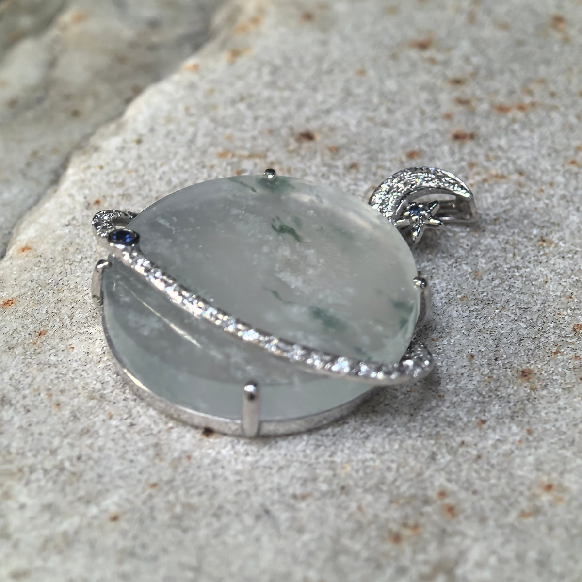 Low angled view of a pale green icy jadeite Planet Pendant on stone, showing its rounded profile, 18K gold setting and diamond-set orbital ring.