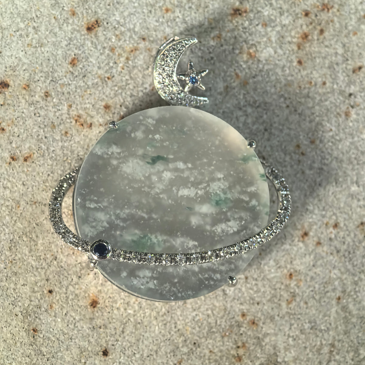 Front view of a pale green jadeite Planet Pendant on textured stone, showing white snowflake-like inclusions, green accents and the diamond-set ring surrounding the center.