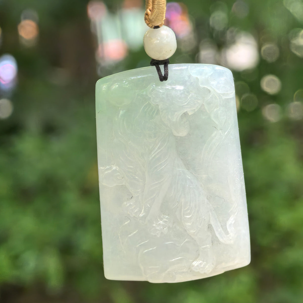 Glutinous pale green jadeite tiger plaque pendant hanging from a cord against a green background, showing the ascending tiger relief.
