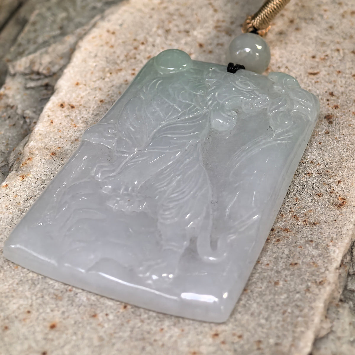 Low angled view of a pale green jadeite tiger plaque pendant on stone, highlighting the relief carving and polished surface.