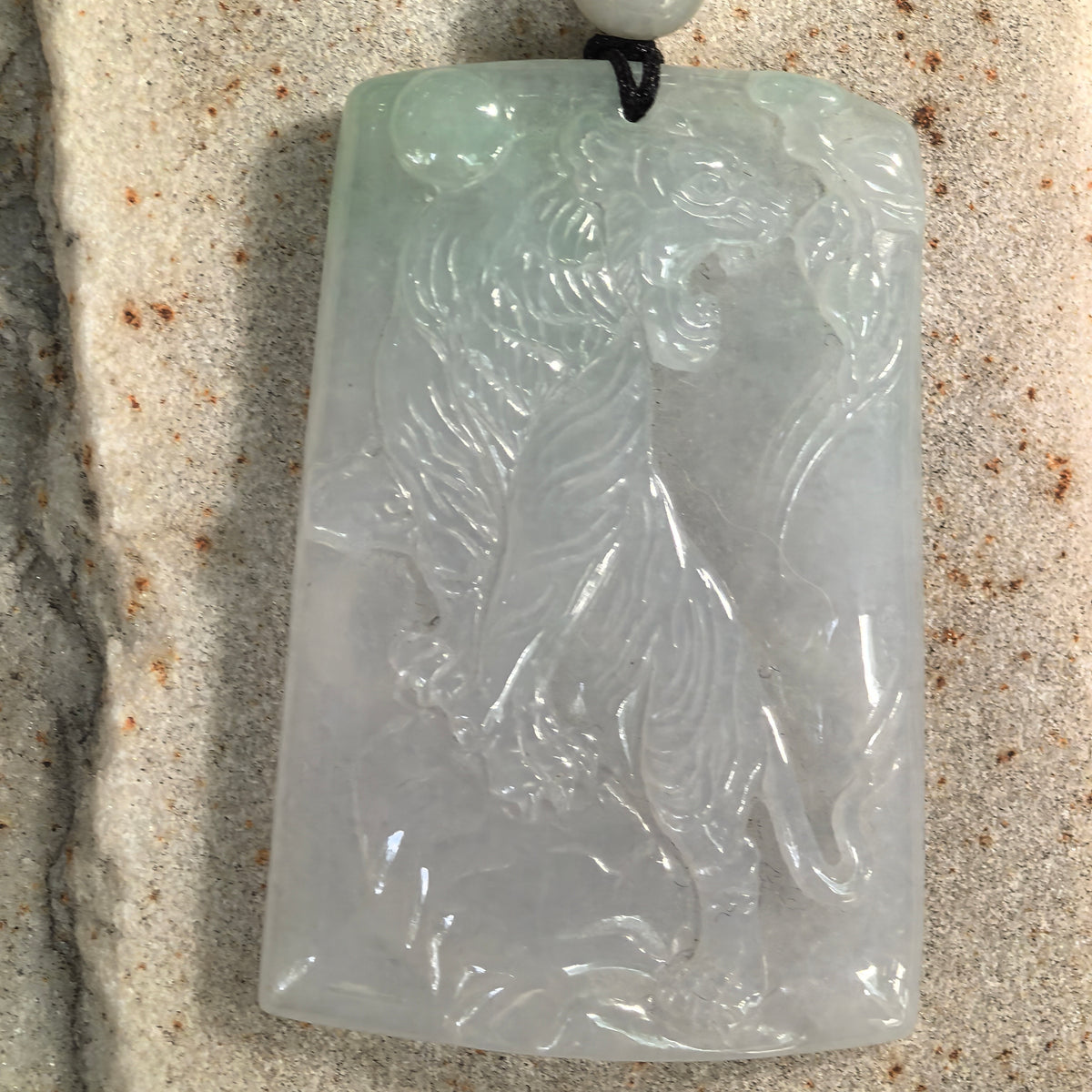 Front close-up of a pale green jadeite tiger plaque pendant on stone, showing the tiger’s raised head, carved stripes and long curved tail.