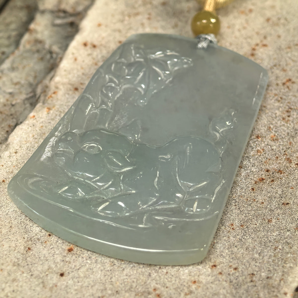 Low angled view of a pale green jadeite Pig Plaque Pendant on stone, highlighting the pig’s defined snout, rounded body and raised tail.