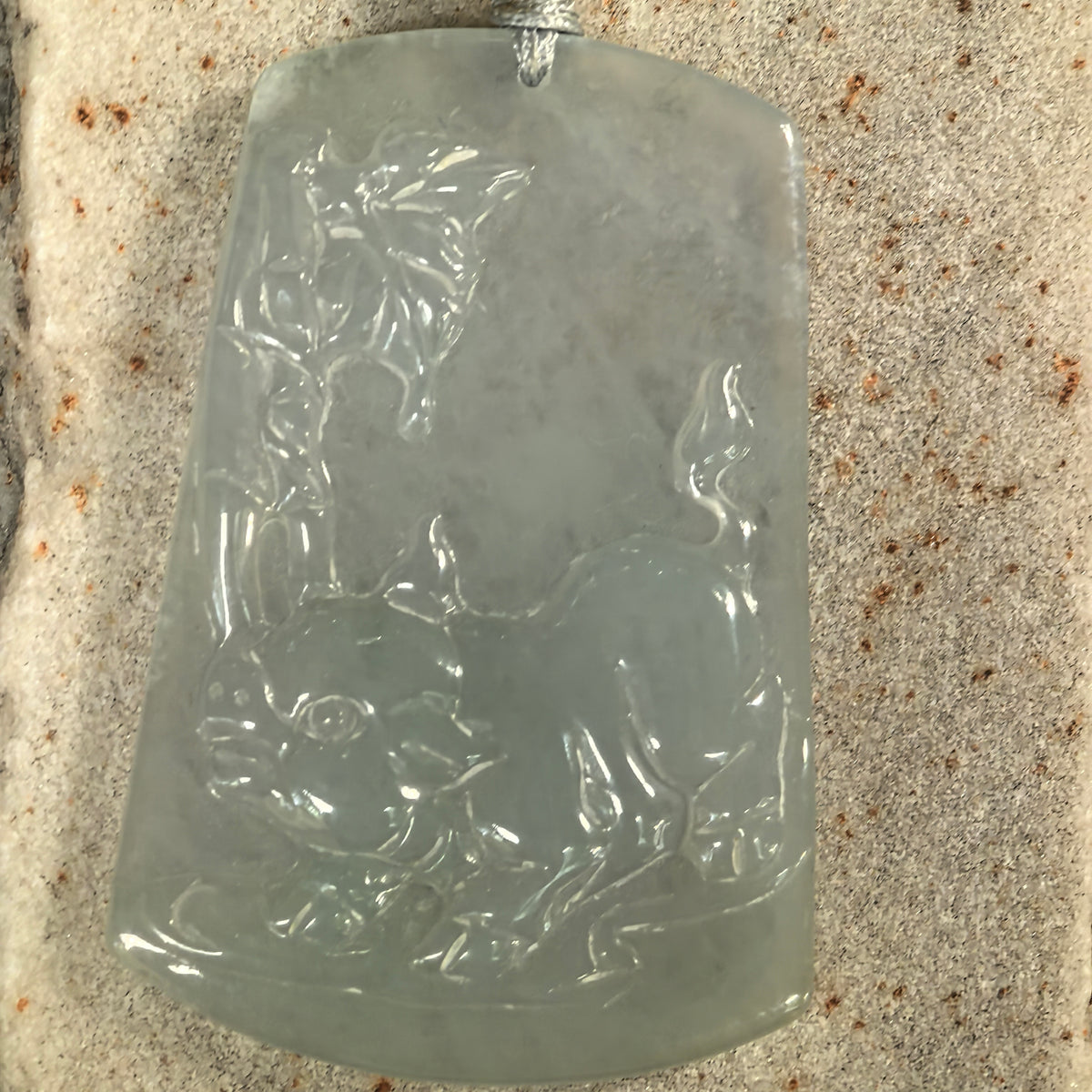 Front view of a pale green jadeite Pig Plaque Pendant on stone, showing the animal relief and leafy carving along one side.
