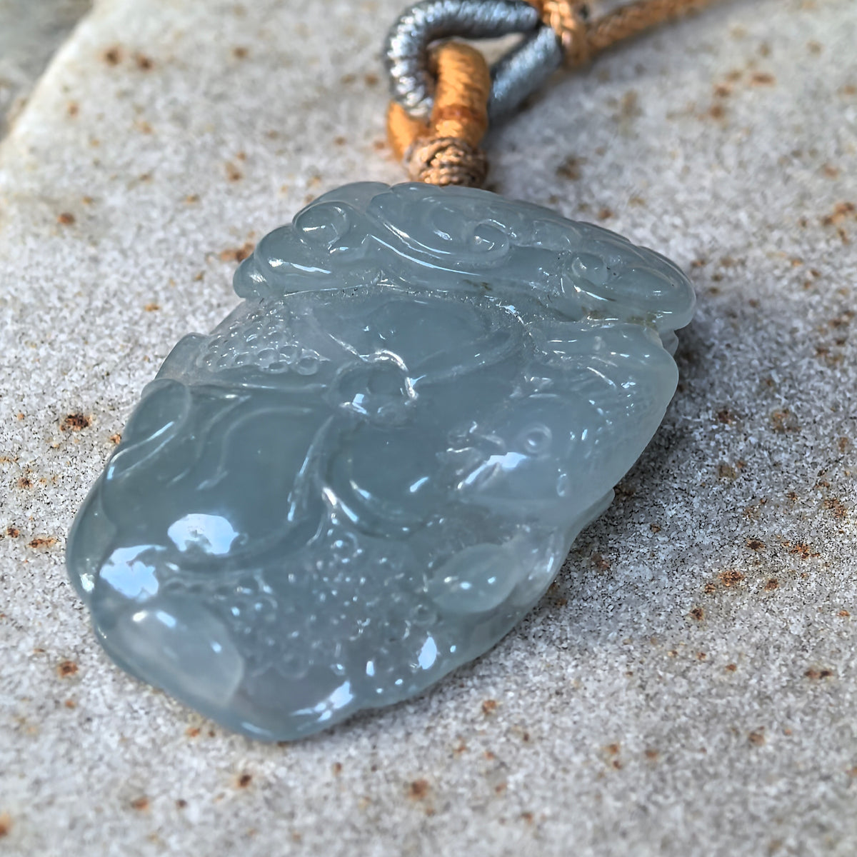 Low angled close-up of a blue lotus jadeite pendant on stone, highlighting the rounded carving, polished surface and thickness.
