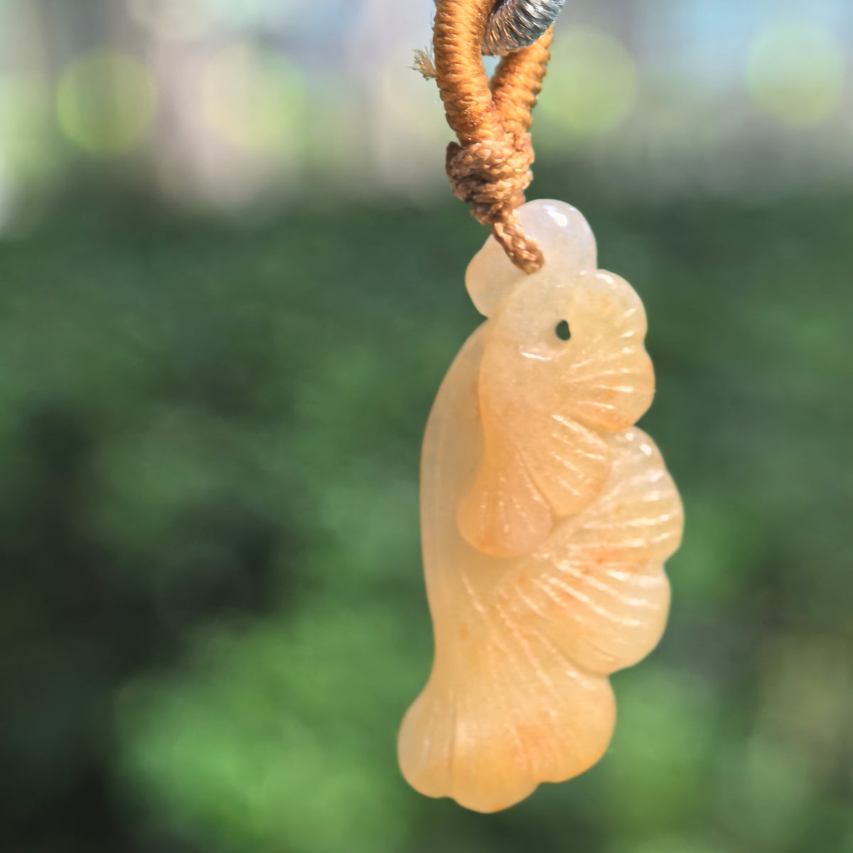 Yellow jadeite ginkgo leaf pendant hanging from a cord against a blurred green background, showing overlapping leaves and radiating carved veins.