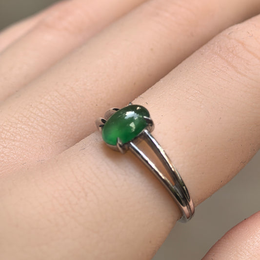 Green oval jadeite ring worn on a finger, showing its glossy cabochon, four platinum prongs and slim split shoulders.