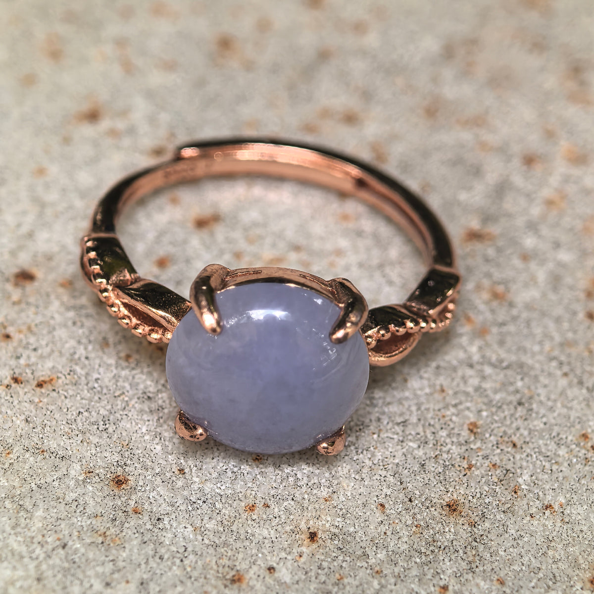 Front view of a nearly round purple jadeite cabochon on a textured stone surface, showing the warm gold-tone prongs and decorative shoulders.