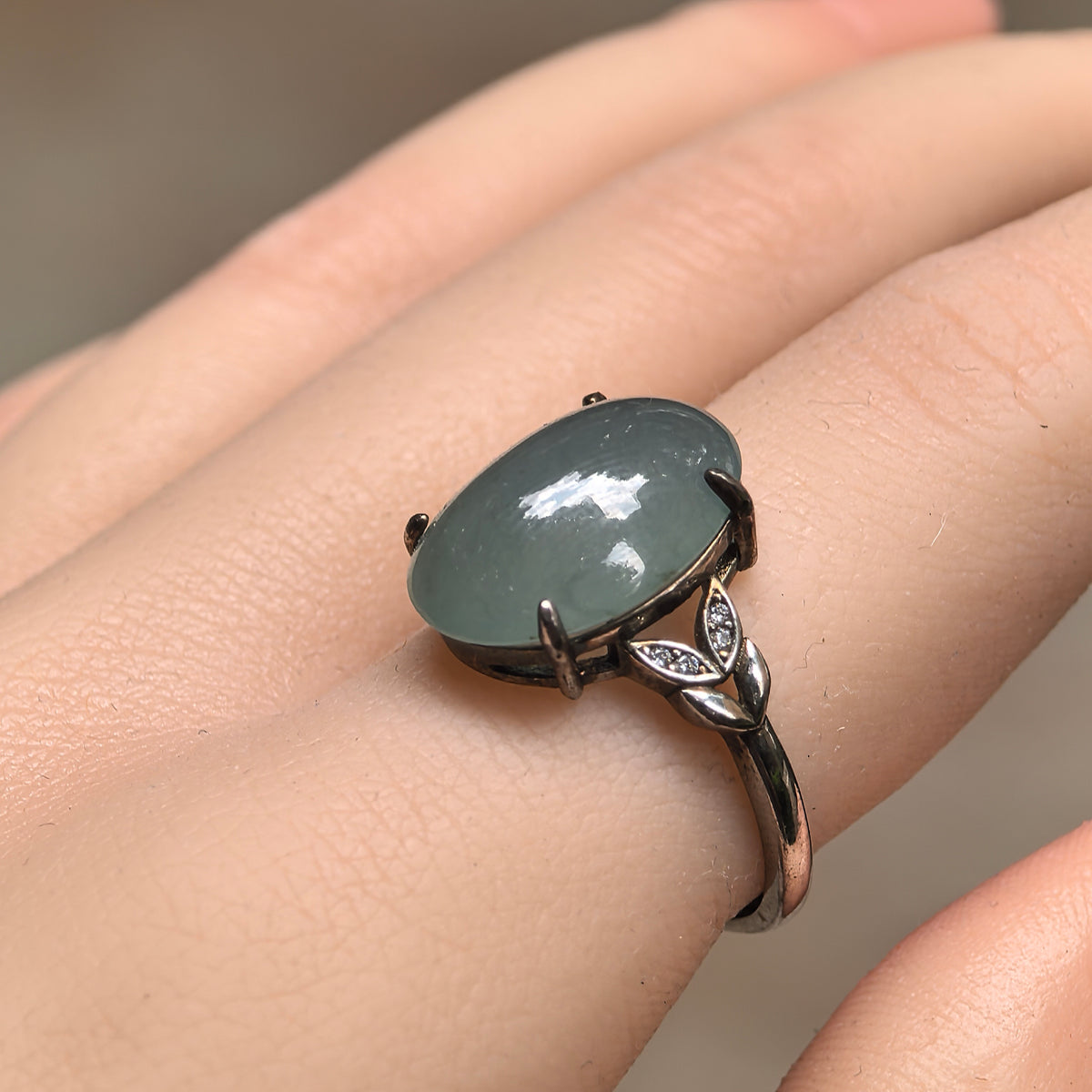 Side view of a lake green oval jadeite ring worn on a finger, showing the domed cabochon and leaf-shaped silver shoulder details with clear accent stones.