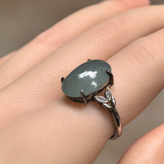 Side view of a lake green oval jadeite ring worn on a finger, showing the domed cabochon and leaf-shaped silver shoulder details with clear accent stones.