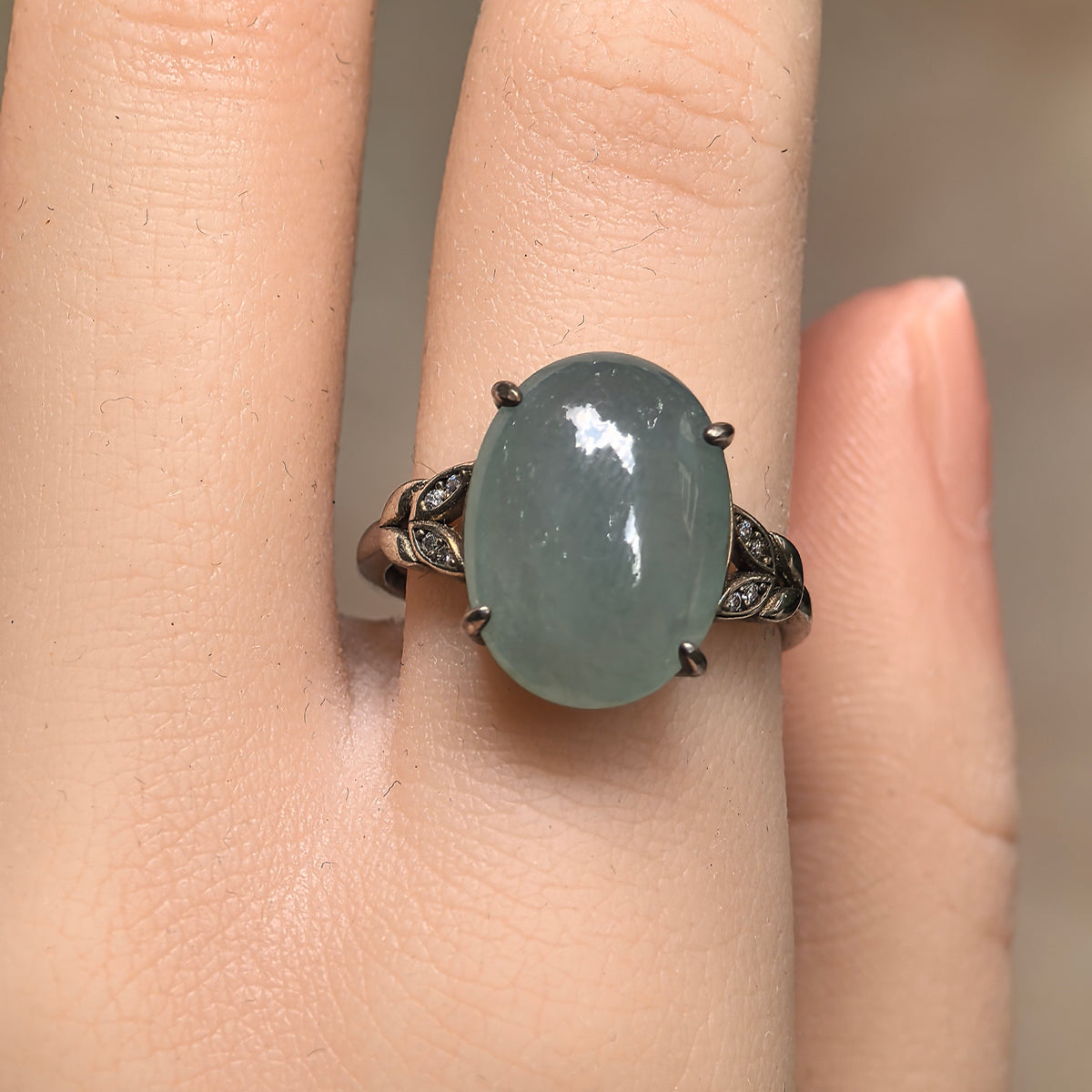 Front view of a lake green oval jadeite ring worn on a finger, showing the four-prong setting and silver leaf details on both shoulders.