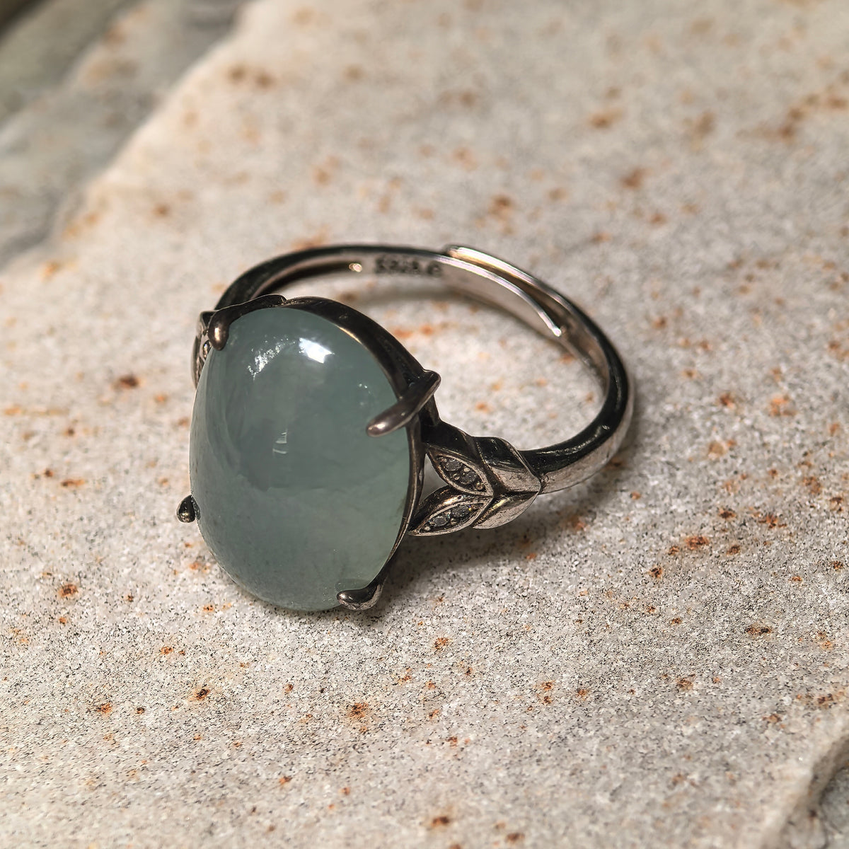 Angled close-up of a lake green jadeite ring on a textured stone surface, showing leaf-shaped shoulder details, clear accent stones and the open silver band.