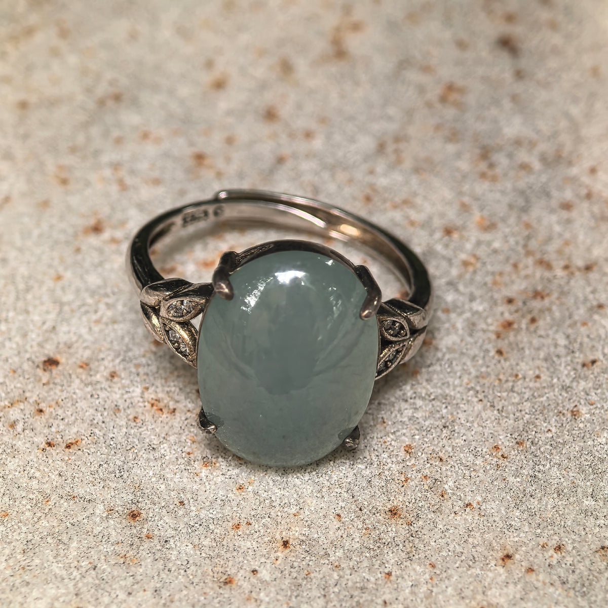 Front close-up of an oval lake green jadeite cabochon in S925 sterling silver, with leaf details beside the stone and the adjustable band visible behind it.