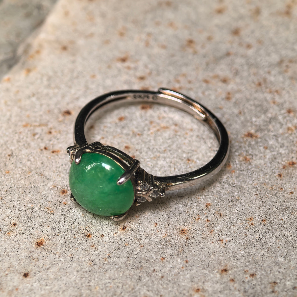 Green round jadeite ring resting on a stone surface, viewed at an angle to show the silver prongs, shoulder accents, and adjustable open band.