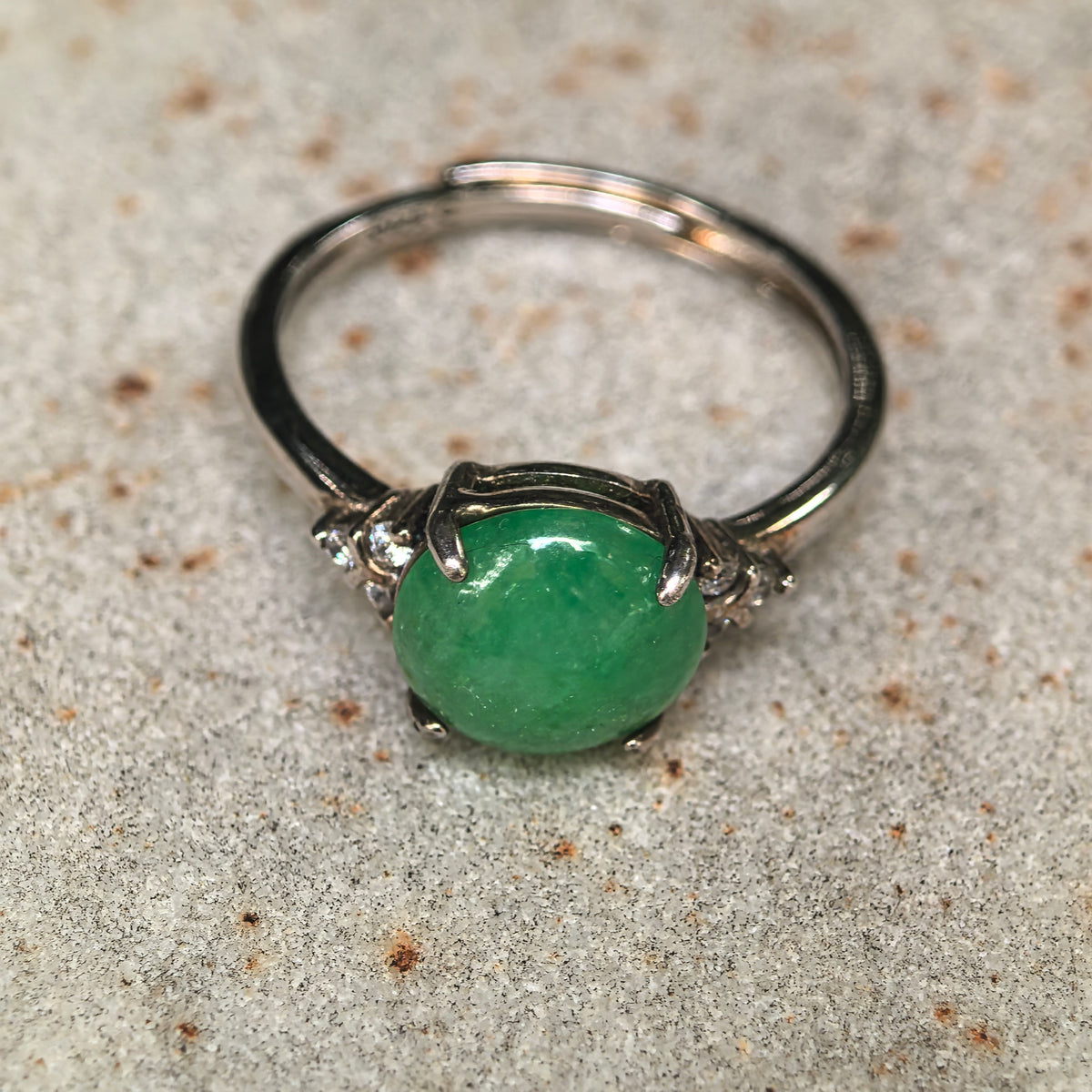 Front close-up of a green jadeite ring on a stone surface, showing the domed cabochon, S925 silver setting, and clear accent stones.