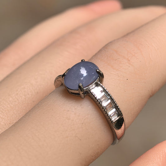 Angled view of a purple oval jadeite ring worn on a finger, showing the S925 silver prongs and rectangular clear accent stones along the shoulder.