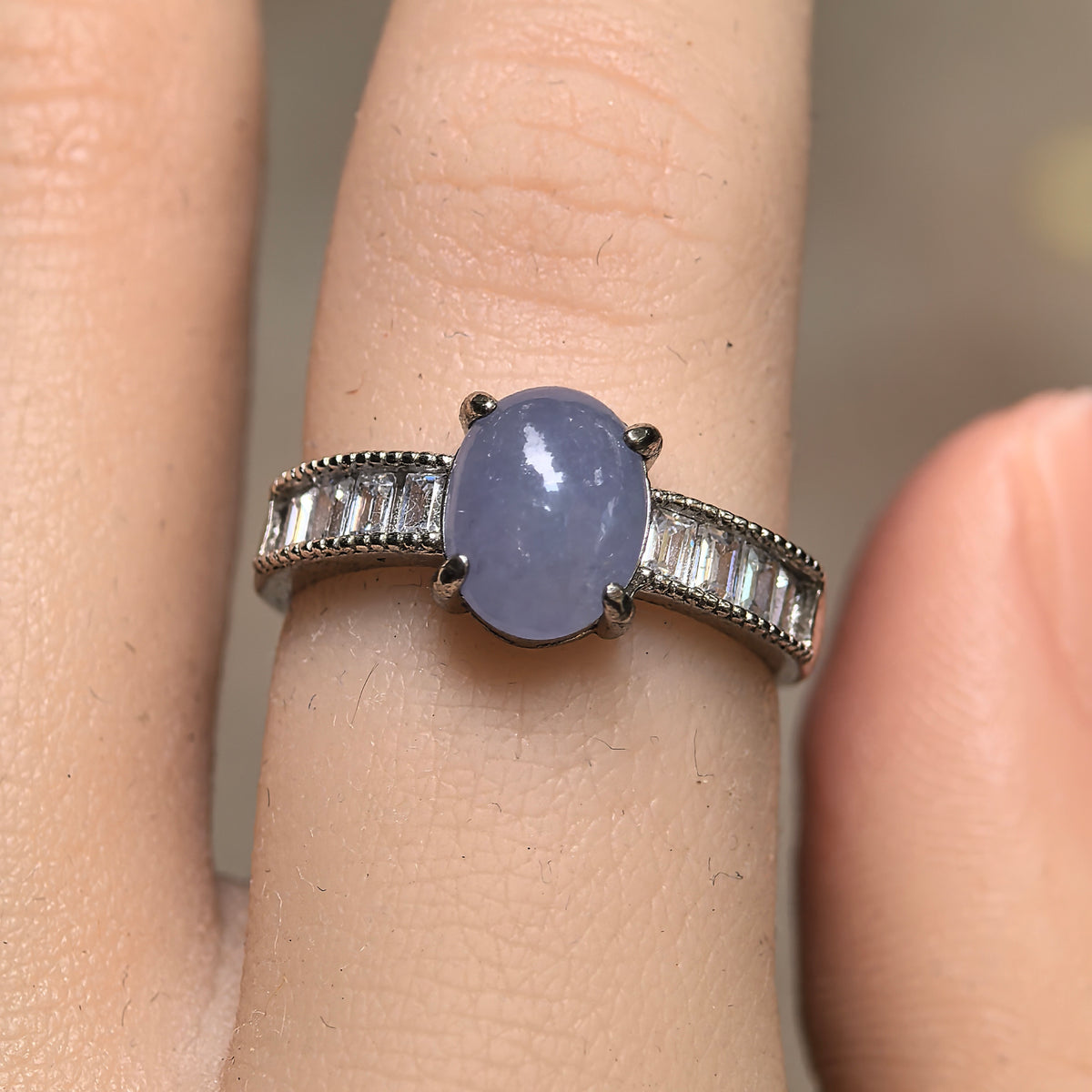 Front view of a glutinous purple jadeite ring worn on a finger, with an oval cabochon and clear accent stones on both silver shoulders.