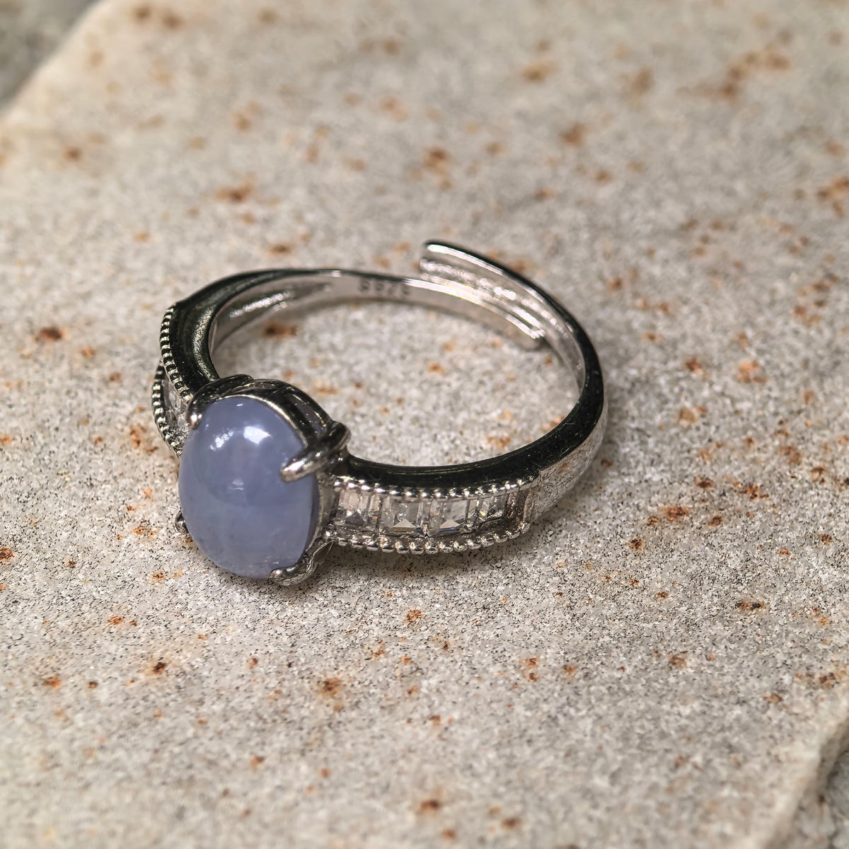 Purple oval jadeite ring resting on a stone surface, showing the clear accent stones, beaded edging, and adjustable open silver band.