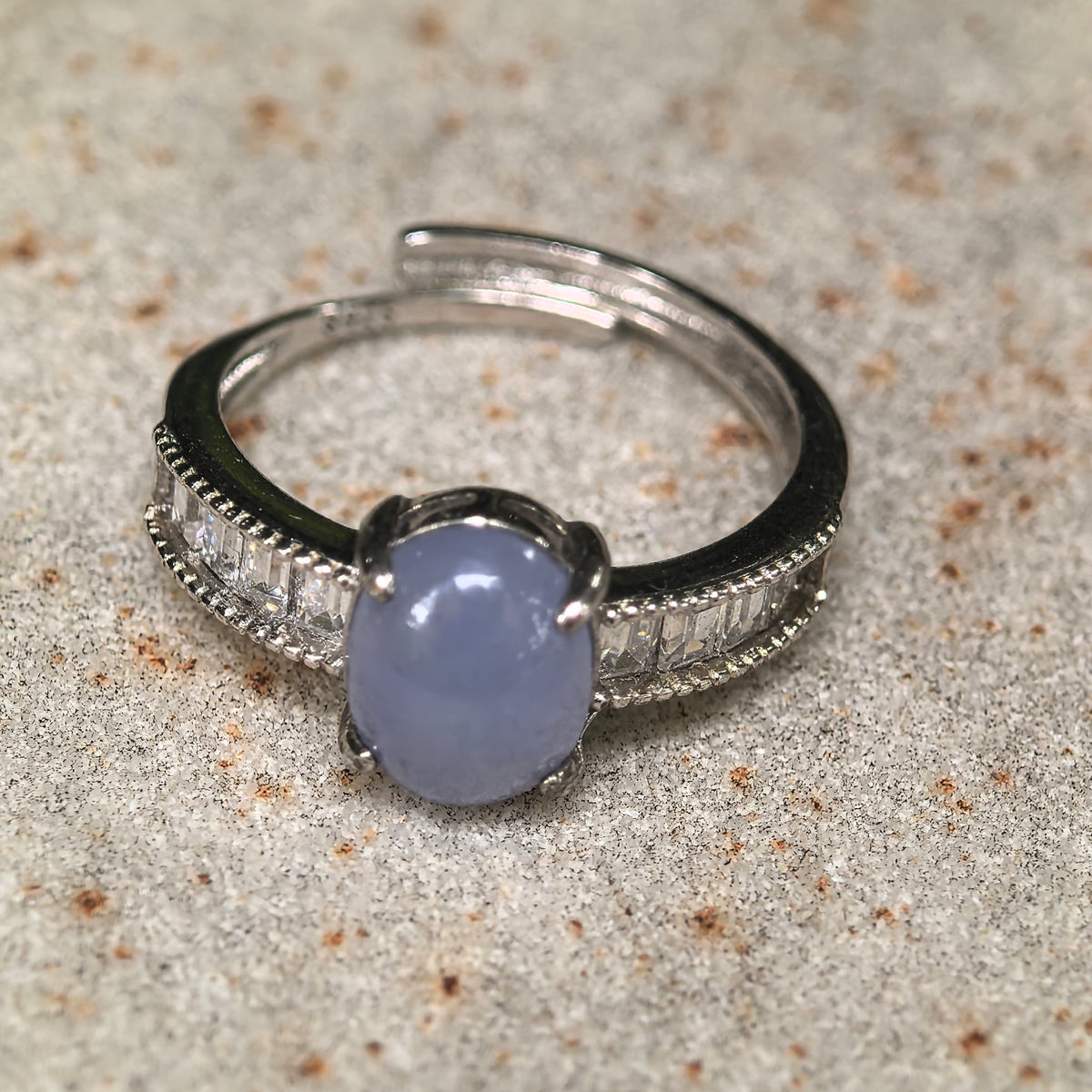 Front close-up of a purple jadeite ring on a stone surface, showing the oval cabochon, S925 silver setting, and rectangular clear accent stones.